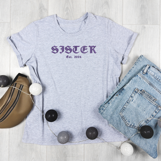 Gray t-shirt with 'SISTER Est. 2024' text, jeans, and a bag on a light wooden surface.