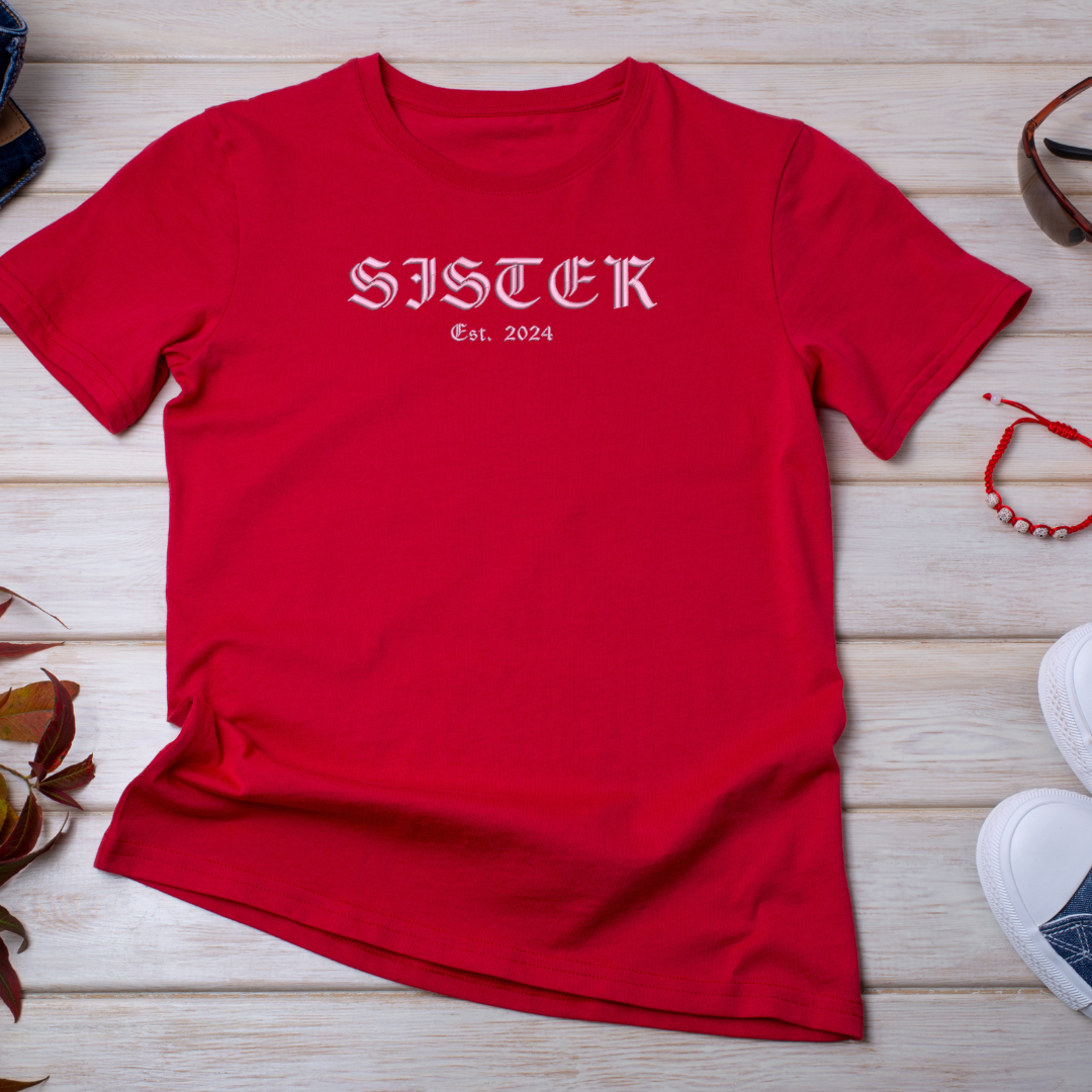 Red t-shirt with 'SISTER Est. 2024' text on a wooden surface with accessories.