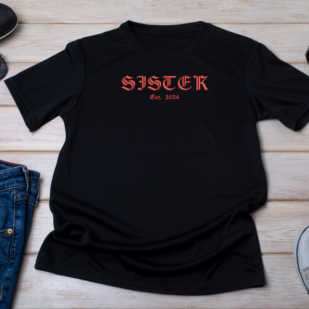 Black t-shirt with 'SISTER Est. 2024' text on a wooden surface with denim jeans.